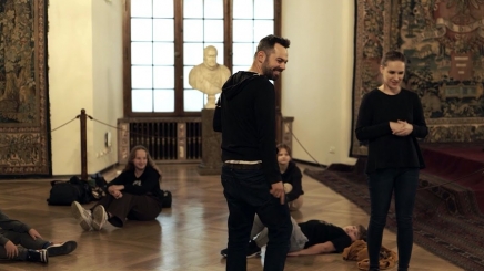 sala w muzeum, kilka osób w czarnych strojach, niektórzy leżą na podłodze a dwie osoby stoją