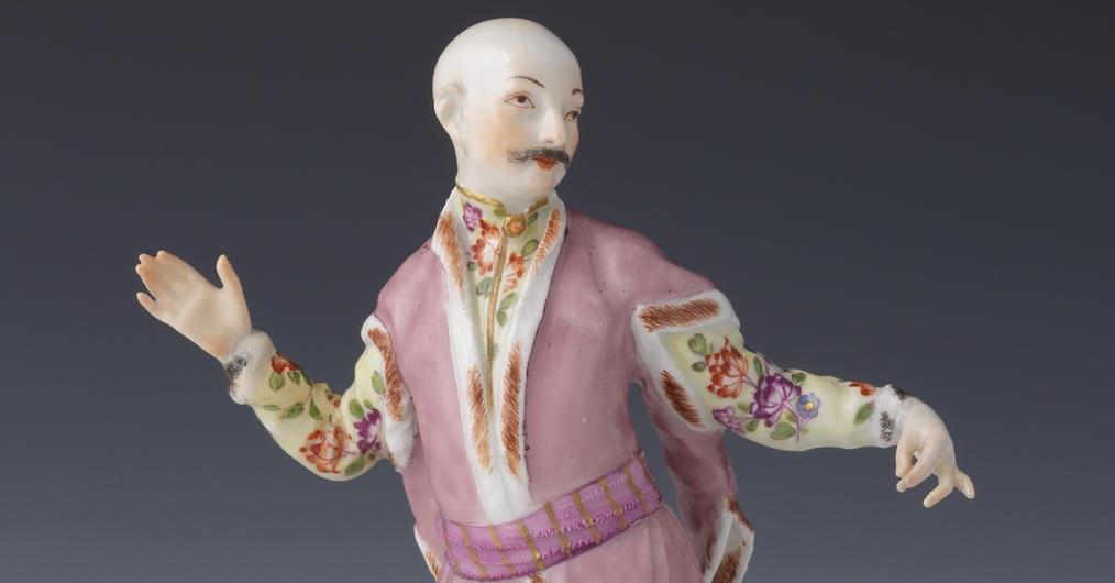 Na zdjęciu fragment porcelanowej figurki z manufaktury w Miśni. Figurka przedstawia łysego mężczyznę z wąsami, ubranego w fioletowy kontusz i koszulę w kolorowe kwiaty. Figurka sfotografowana jest od pasa w górę i umieszczona na szarym tle.