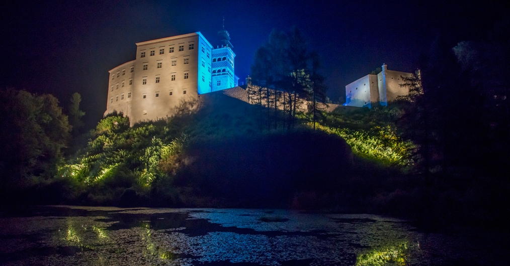 Zdjęcie przedstawia Zamek w Pieskowej Skale widziany od podnóża góry. Budynek jest oświetlony żółtym i niebieskim światłem, otoczony trawą i drzewami.