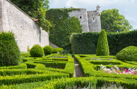 Zdjęcie przedstawia zielony ogród zamkowy. Pod jasnoszarymi murami rozciąga się przystrzyżony bukszpan oraz krzewy.