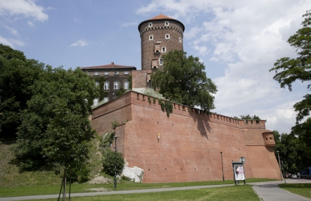 Na zdjęciu widać Wawel od strony Wisły - ceglany mur, basztę i fragment zamku w otoczeniu zielonych drzew.