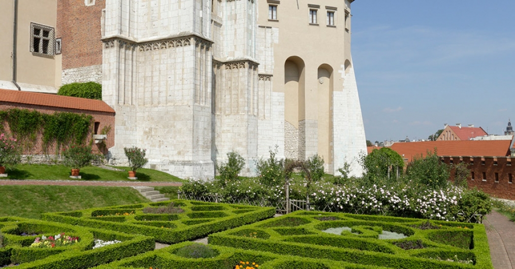 Ogrody królewskie, zielone rabaty, kwitnące rośliny, po lewej stronie fragment ściany Zamku, w tle niebo