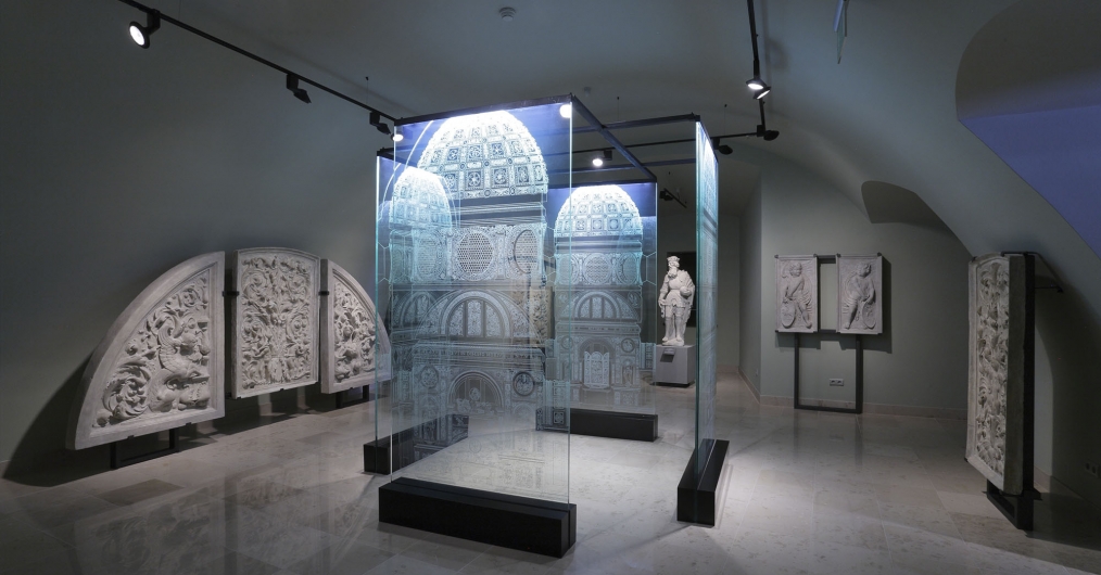 Zdjęcie przedstawia salę wystawową. Pod ścianami ustawione są płaskorzeźby, a na środku stoi instalacja ze szklanych paneli, na których ukazana jest fasada kaplicy.