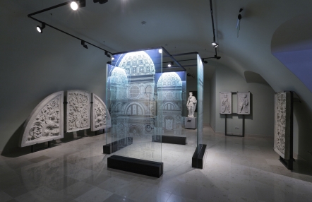 Zdjęcie przedstawia salę wystawową. Pod ścianami ustawione są płaskorzeźby, a na środku stoi instalacja ze szklanych paneli, na których ukazana jest fasada kaplicy.
