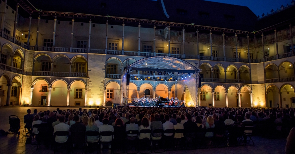 Zdjęcie przedstawia występ orkiestry na wieczornym, oświetlonym dziedzińcu arkadowym Wawelu.