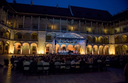 Zdjęcie przedstawia występ orkiestyry na wieczornym, oświetlonym dziedzińcu arkadowym wawelu.