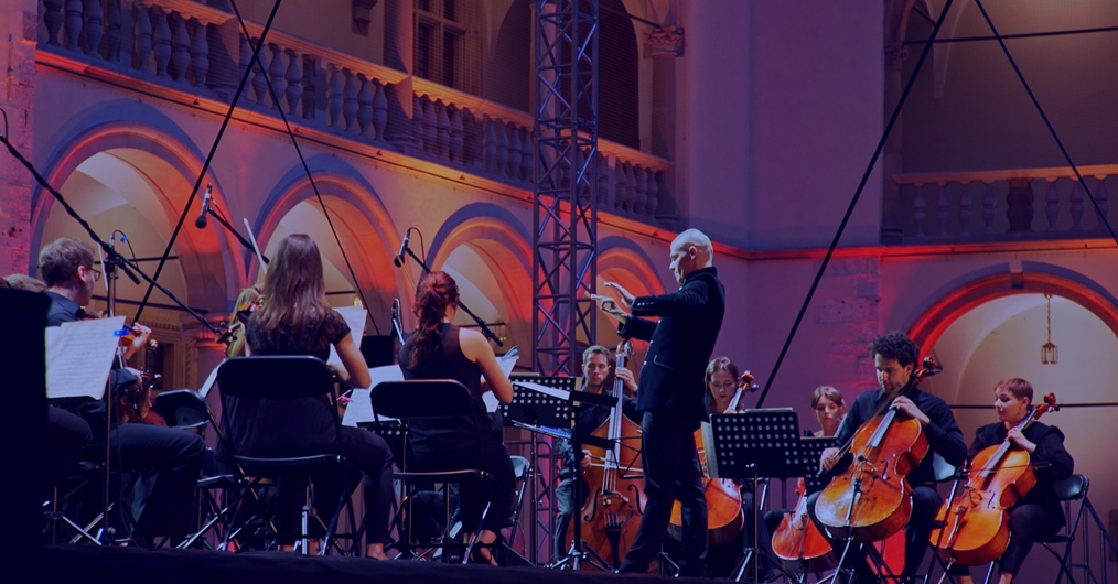 grupa muzyków grających na instrumentach smyczkowych, z  prawej strony postać dyrygenta, w tle krużganki wawelskiego  zamku