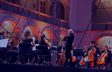 grupa muzyków grających na instrumentach smyczkowych, z  prawej strony postać dyrygenta, w tle krużganki wawelskiego  zamku