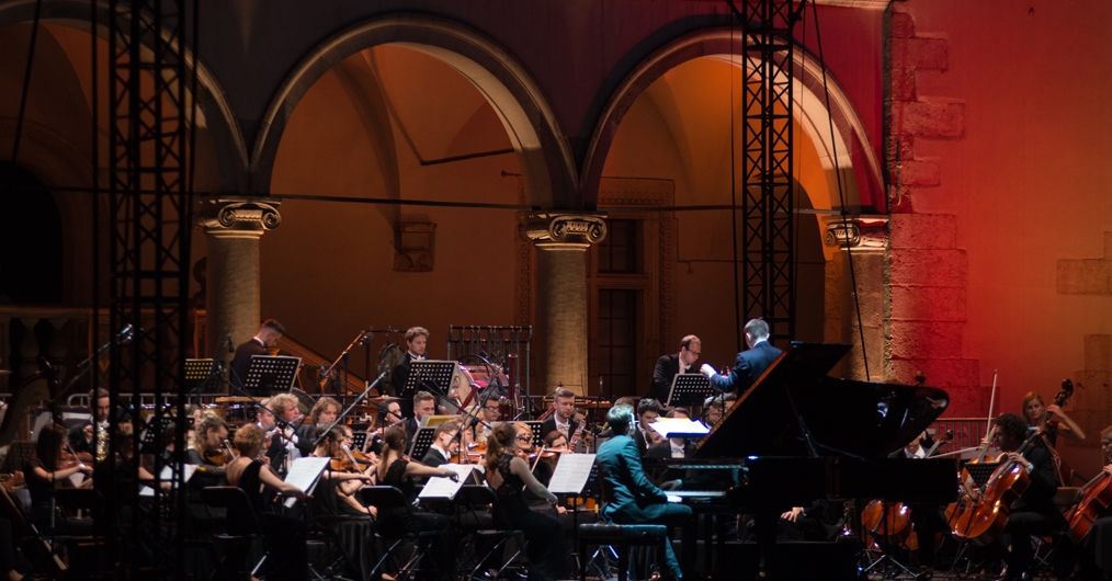 fragment dziedzińca arkadowego  wawelskiego zamku, podświetlony nastrojowym światłem,  na scenie dyrygent i muzycy grający na instrumentach smyczkowych oraz pianista przy fortepianie