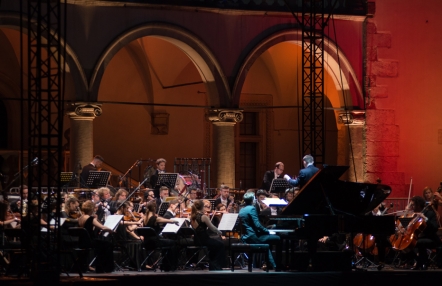 fragment dziedzińca arkadowego  wawelskiego zamku, podświetlony nastrojowym światłem,  na scenie dyrygent i muzycy grający na instrumentach smyczkowych oraz pianista przy fortepianie