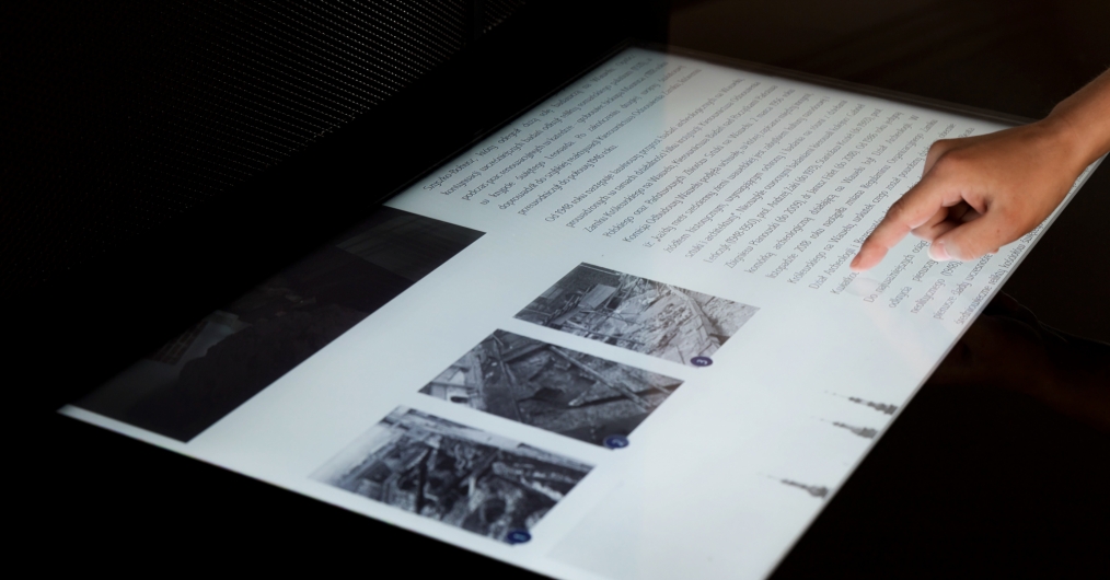 Ekran dotykowy z fragmentem tekstów i zdjęciami, z prawej strony ręka z palcem dotykającym ekran
