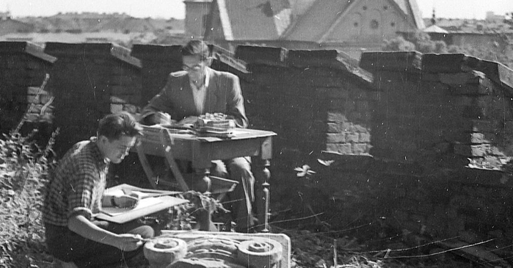 Widok zewnętrzny, przy stoliku piszący mężczyzna, przed nim mężczyzna siedzący na trawie, na kolanach ma deskę rysunkową, po prawej stronie fragment głowicy zabytkowej kolumny, w tle mur