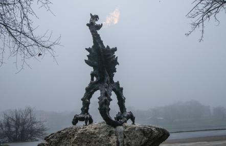 rzeźba dużego stojącego smoka, w tle zamglony krajobraz; z pyska smoka wydobywa się ogień