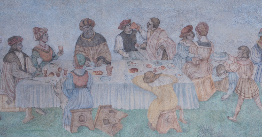 fragment naściennego malowidła, przedstawia grupę ludzi w dawnych strojach, siedzą za stołem, jedzą, rozmawiają