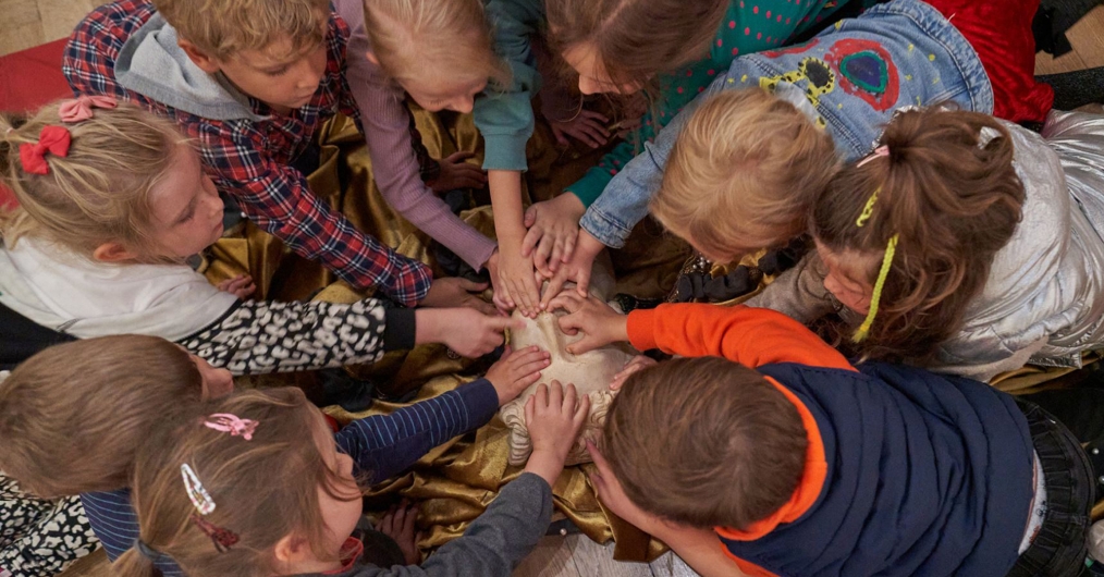 Grupa dzieci w wieku szkolnym siedzi na podłodze, tworząc krąg nad tkaniną