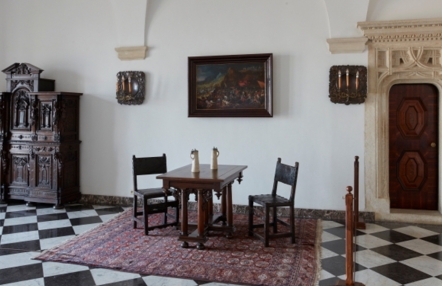 komnata w zamku, z lewej strony renesansowy portal, na dywanie ustawiony zabytkowy stół i dwa krzesła, na ścianie obracy i kandelabry, z lewej strony bogato rzeźbiona zabytkowa szafa