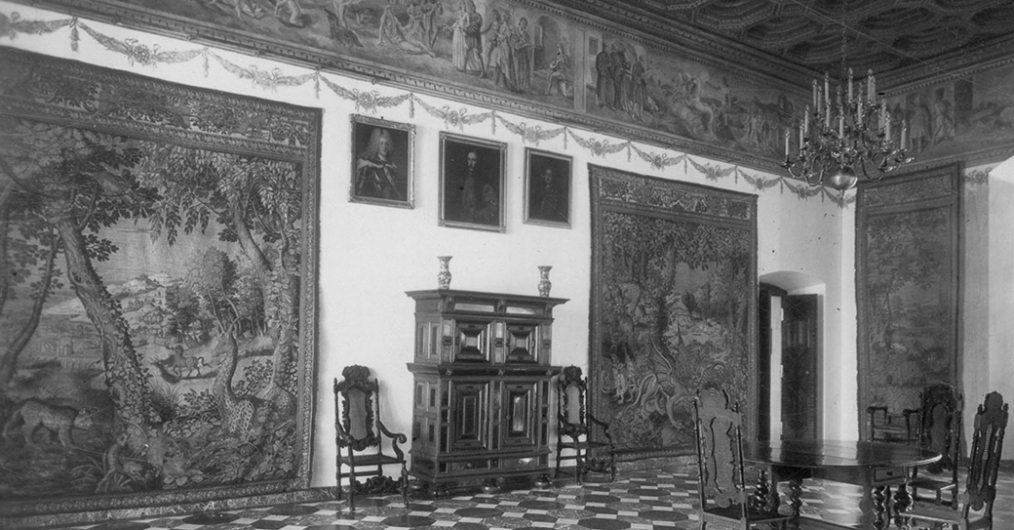 czarno-białe zdjęcie, zabytkowa sala zamku, na ścianie po lewej stronie obrazy i tkaniny, między nimi zabytkowe meble - szafa i dwa krzesła, pod sufitem malowidło okalające salę, z prawej strony okrągły stół i krzesła