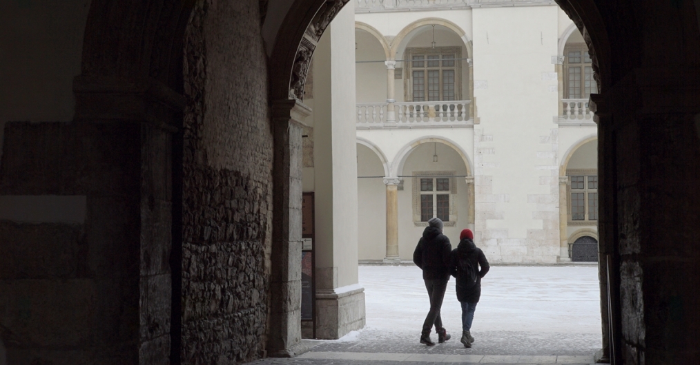 Dwoje ludzi przechodzi przez bramę, w tle widok dziedzińca arkadowego na Wawelu.