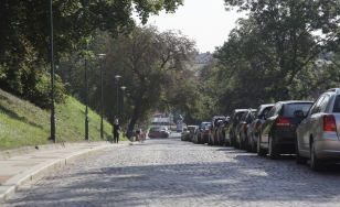 stromy zjazd w dół, nierówna kostka brukowa na jezdni, nierówne płyty chodnikowe z wysokim progiem, wzdłuż drogi po prawej stronie stoją samochody