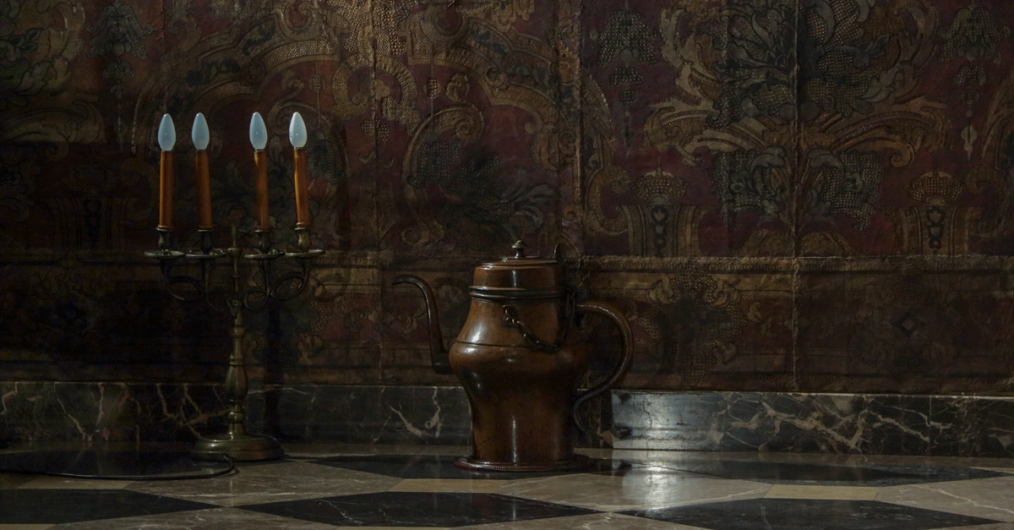 fragment ciemnej sali, widać fragment ściany i podłogi, ściana pokryta materiałem z ornamentami, na podłodze, po lewej stronie stoi świecznik z czterema światłami, pośrodku zabytkowy miedziany dzbanek