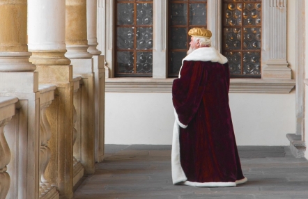 mężczyzna w czerwonej szacie obszytej białym futrem i złotej czapce idzie przez krużganki wawelskiego zamku