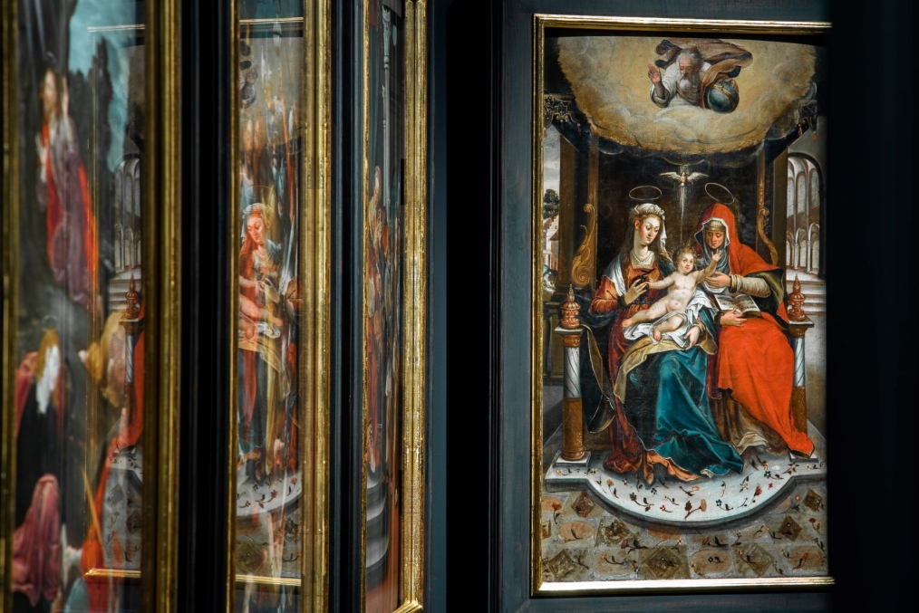 z lewej strony fragmenty religijnych obrazów, w tle obraz z Matką Boską, któa na kolanach trzyma Dzieciątko; z pawej strony siedząca postać kobieca w czerwonej szacie