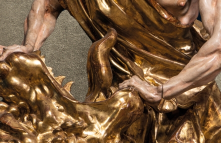 fragment rokowej drewnianej rzeźby; umięśnione ręce mężczyzny oparte o bogato rzeźbiony i złocony element