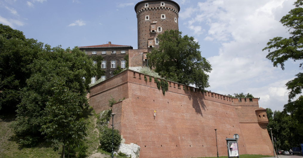 Na zdjęciu widać Wawel od strony Wisły - ceglany mur, basztę i fragment zamku w otoczeniu zielonych drzew.
