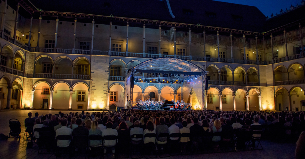 Wydarzenie muzyczne na wieczornym dziedzińcu arkadowym. Na zdjęciu widać oświetloną scenę i widownię.