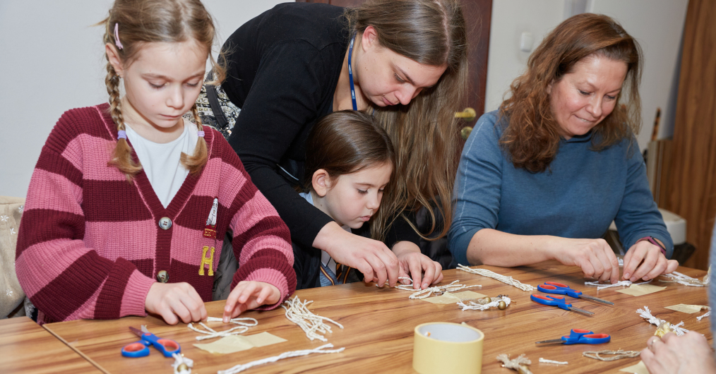 Kobiety i dzieci zaangażowane w twórczą pracę, otoczone kolorowymi materiałami i narzędziami.