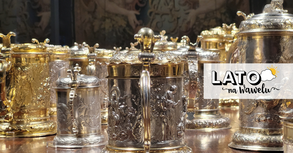 złote i srebrne kubki i kufle, zdobione puncowanymi motywami figuralnymi, w tle fragment arrasu z monogramem króla Zygmunta Augusta (SA)