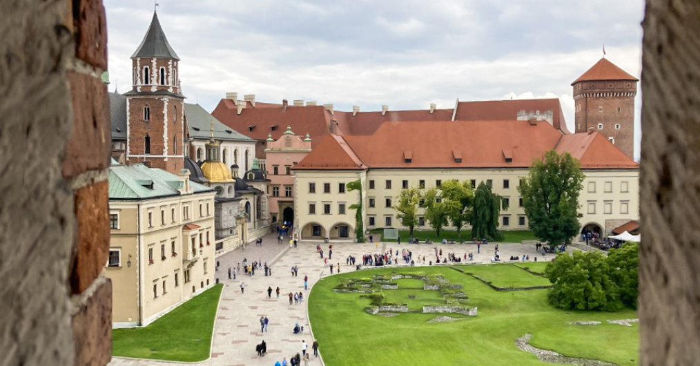 widok z okna baszty na dziedziniec zewnętrzny zamku, po lewej stronie katedra, na wprost budynek administracyjny zamku, w dole na dziedzińcu grupki spacerujących ludzi; letni, nieco pochmurny dzień