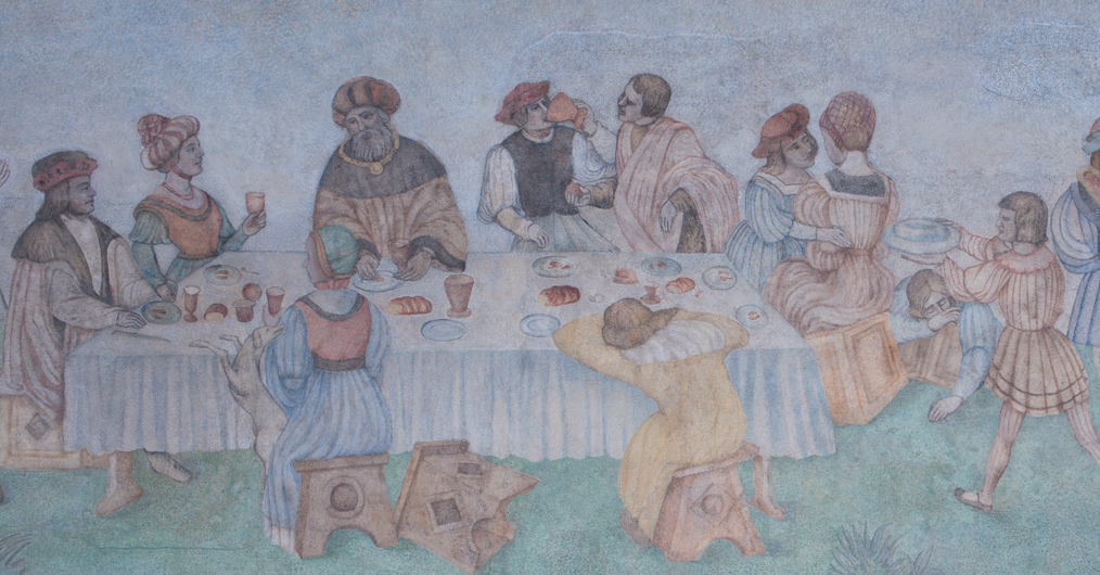 fragment naściennego malowidła, przedstawia grupę ludzi w dawnych strojach, siedzą za stołem, jedzą, rozmawiają