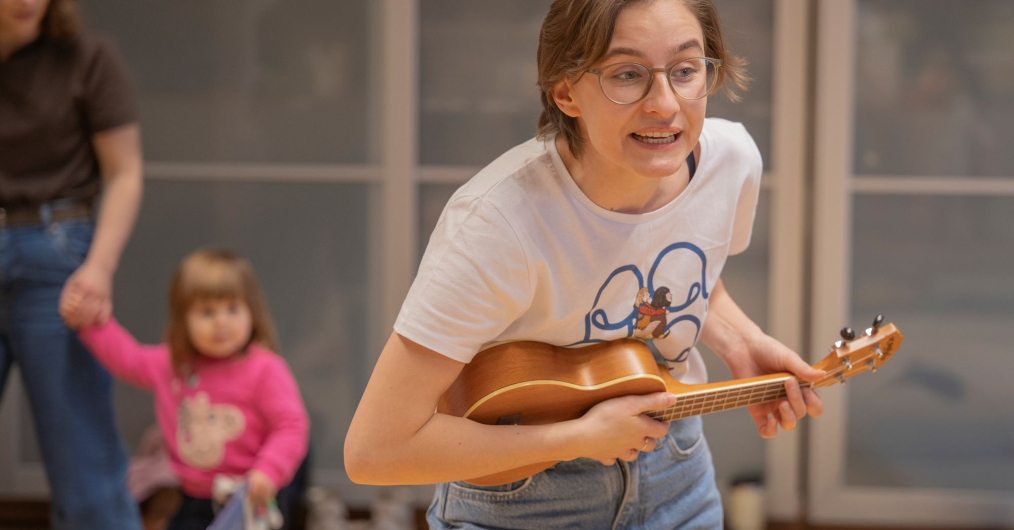 Wesoła scena: kobieta gra na ukulele w towarzystwie innych ludzi w przytulnym pomieszczeniu.