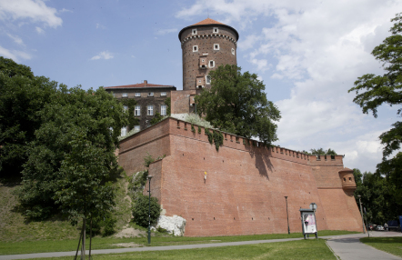 Na zdjęciu widać Wawel od strony Wisły - ceglany mur, basztę i fragment zamku w otoczeniu zielonych drzew.