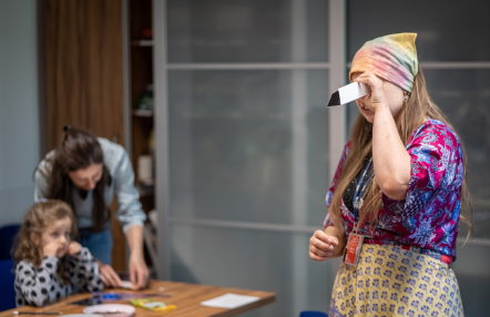 Kobieta ubrana kolorowo patzry przez papierową lunetę, z tyłu przy stole dziewczynka z opiekunką