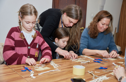 Kobiety i dzieci zaangażowane w twórczą pracę, otoczone kolorowymi materiałami i narzędziami.