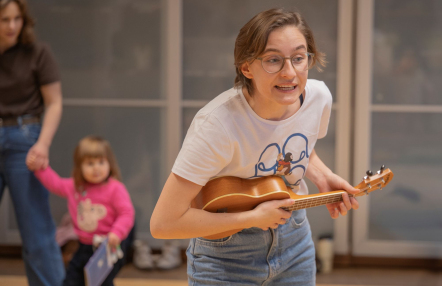 Wesoła scena: kobieta gra na ukulele w towarzystwie innych ludzi w przytulnym pomieszczeniu.