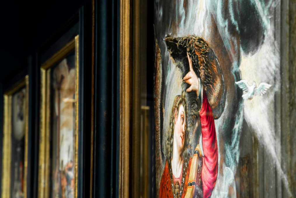 fragment obrazu z postacią anioła, który podnosi do góry lewą rękę, w tle inne obrazy w złoconych ramach