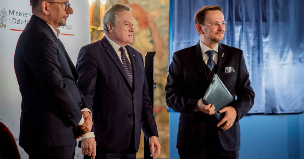 trzech mężczyzn ubranych w garnitury, stoją w zabytkowej sali zamku