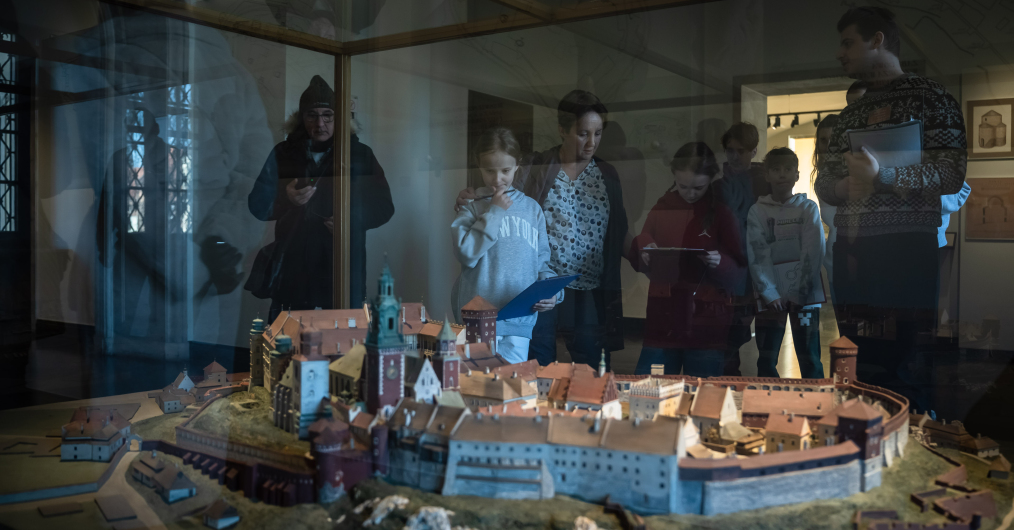 Osoby oglądające wnętrza w muzeum, z zainteresowaniem podziwiające szczegóły wystawy i wypełniające karty zadań.