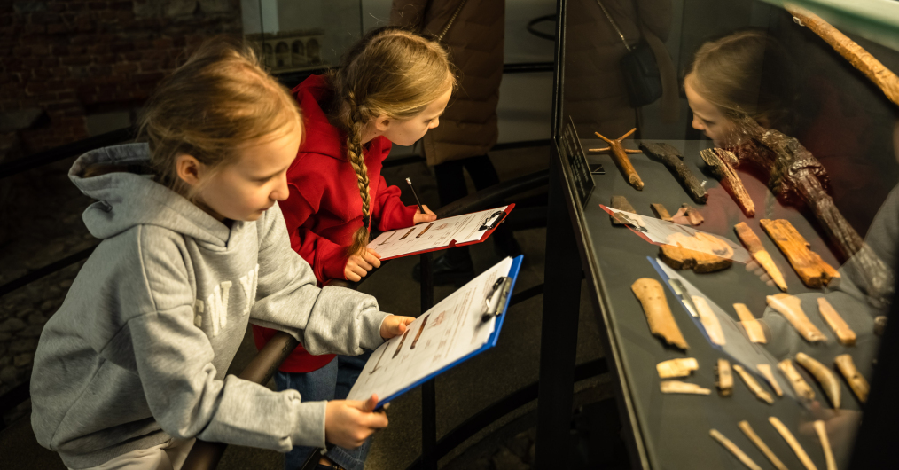 Osoby oglądające wnętrza w muzeum, z zainteresowaniem podziwiające szczegóły wystawy i wypełniające karty zadań.