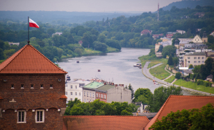 widok na zakole Wisły, budynki i wzgórze porośnięte lasem, na pierwszym planie dachy zamku i biało-czerwona flaga na wieży