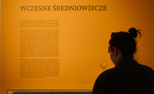 sala wystawy, na ścianie tekst opisowy, w szklanej gablocie obiekty archeologiczne, po prawej stronie sylwetka kobiety stojącej tyłem; ciepłe nastrojowe światło