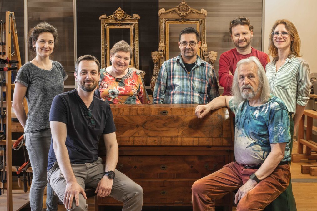 pracownia konserwacji ram, naścianie wisi kilkanaście pustych ram, jedna na drugiej, dwie postacie - kobieta i mężczyzna, pracują przy ramie leżącej na stole