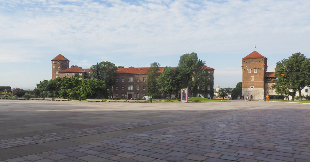 dziedziniec zewnętrzny, duży ceglany budynek nakryty czerwonym dachem, z lewej strony baszta ze spadzistym czerwonym dachem, z prawej strony stojąca oddzielnie baszta ze spadzistym czerwonym dachem