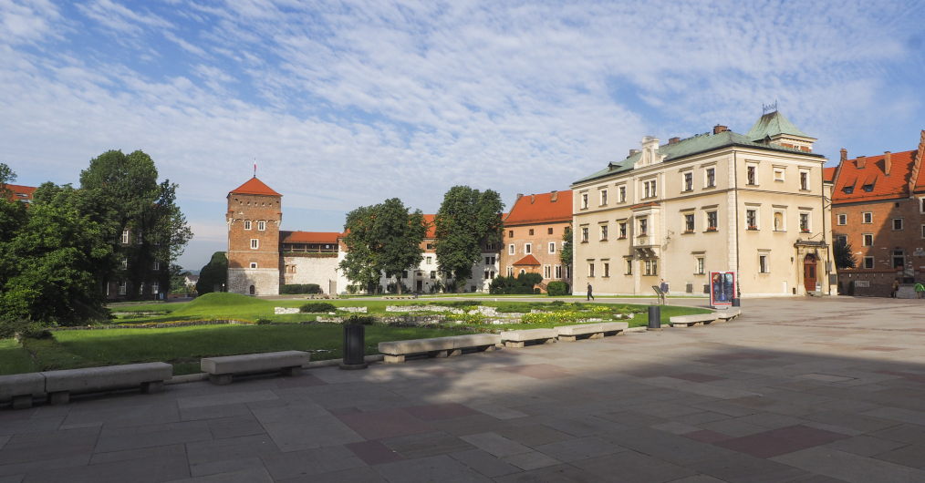 dziedziniec zewnętrzny, klomb, ławki kamienne, po prawej stronie duży jasny budynek, w lewo inne budynki, nakryte czerwonym dachem, po lewej baszta nakryta spadzistym dachem