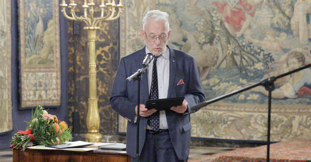 zabytkowa sala zamku, dwie męsie postacie stoją przed mikrofonami, w tle na ścianie arras, po lewej stronie stół z dokumentami i bukietem kwiatów