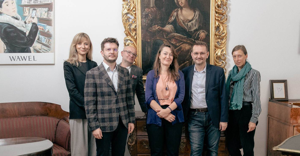 grupa osób we wnętrzu, na ścianie obraz i plakat