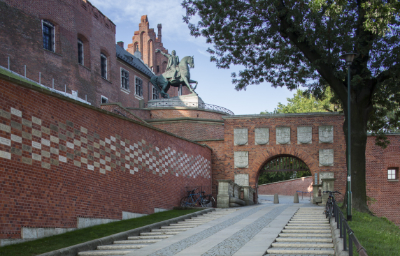 droga prowadząca do bramy, pod górę, z lewej strony ceglany mur z wbudowanymi cegiełkami kamiennymi, nad nim fragment ściany budynku katedry, pomnik mężczyzny siedzącego na koniu, z prawej strony bramy drzewo z zielonymi liśćmi